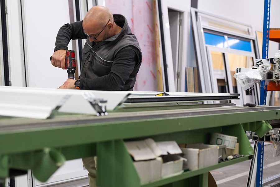 Carpenter using cordless drill assembling an aluminum window frame in a busy workshop, showcasing craftsmanship and industrial manufacturing